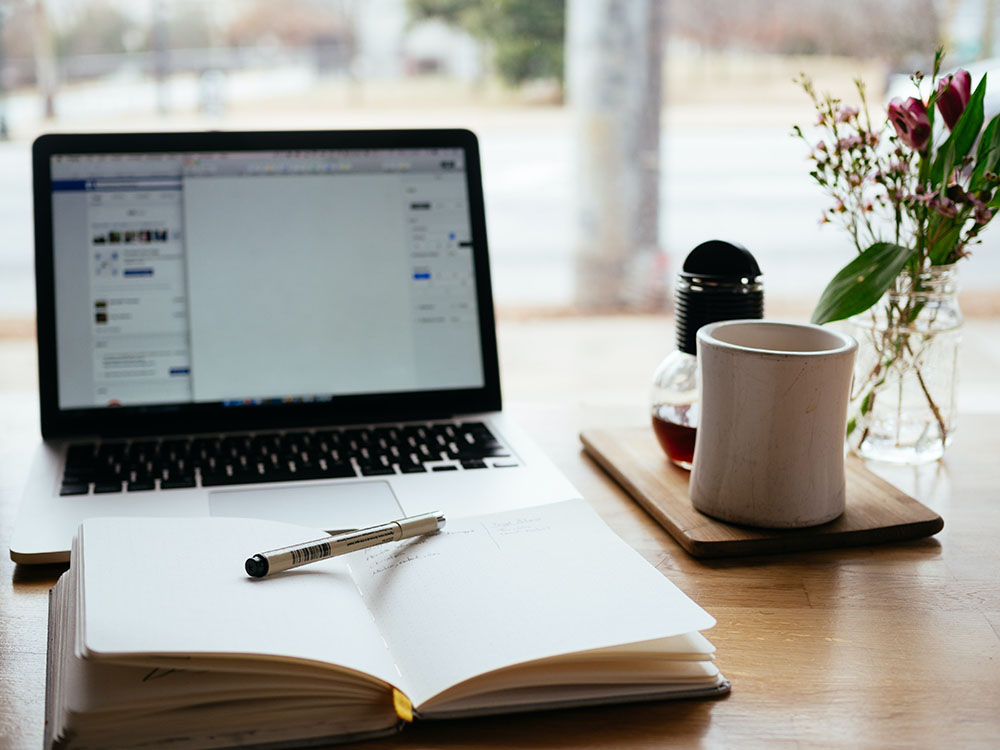 Computer on desk in front of window with notebook, pen and mug